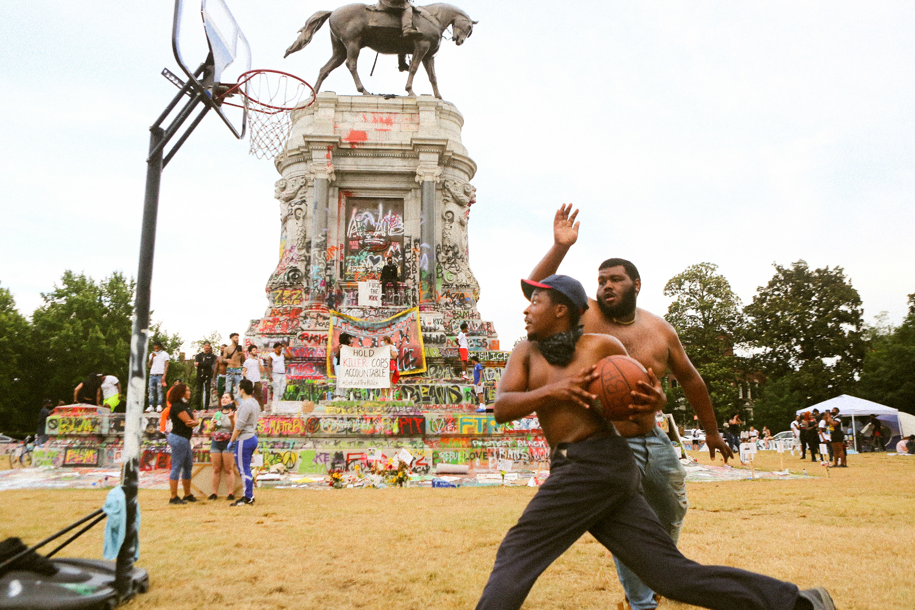 Two men play basketball in the foreground, in the background is a confederate monument covered in graffiti 