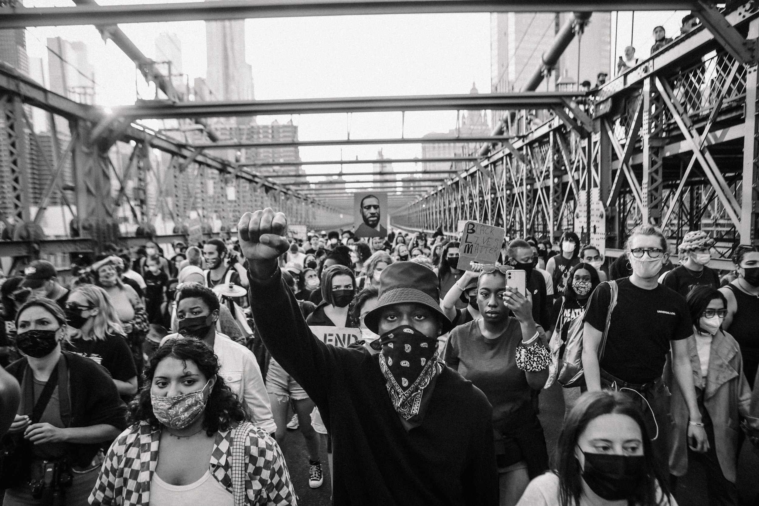 A crowd of protestors wearing masks and with their phones and fists up cross the Brooklyn Bridge in New York