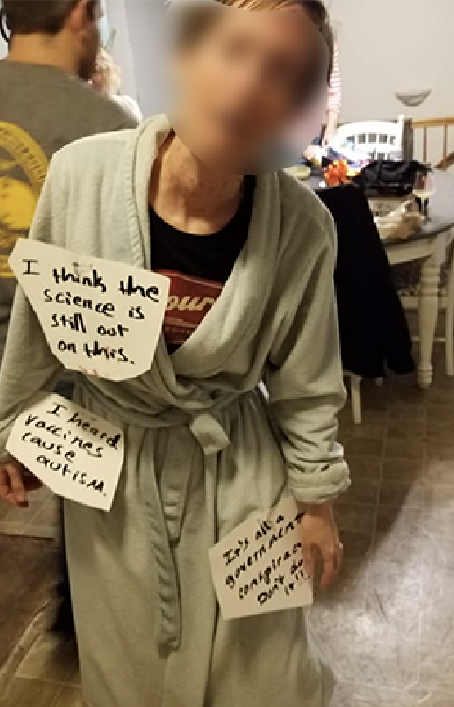 woman in anti-vaxxer costume with a bathrobe and signs saying &quot;I heard vaccines cause autism&quot; and other anti-vaxxer sentiments