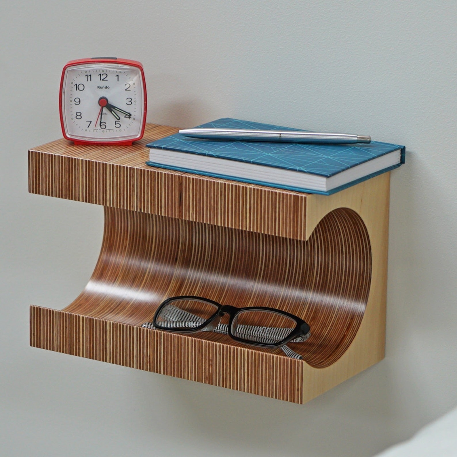a floating nightstand with two tiers, holding an alarm clock, notebook, and pair of glasses