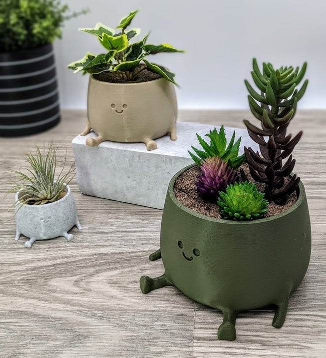 Product image of small gray smiling pot, brown medium smiling pot, and large dark green smiling pot with plants inside