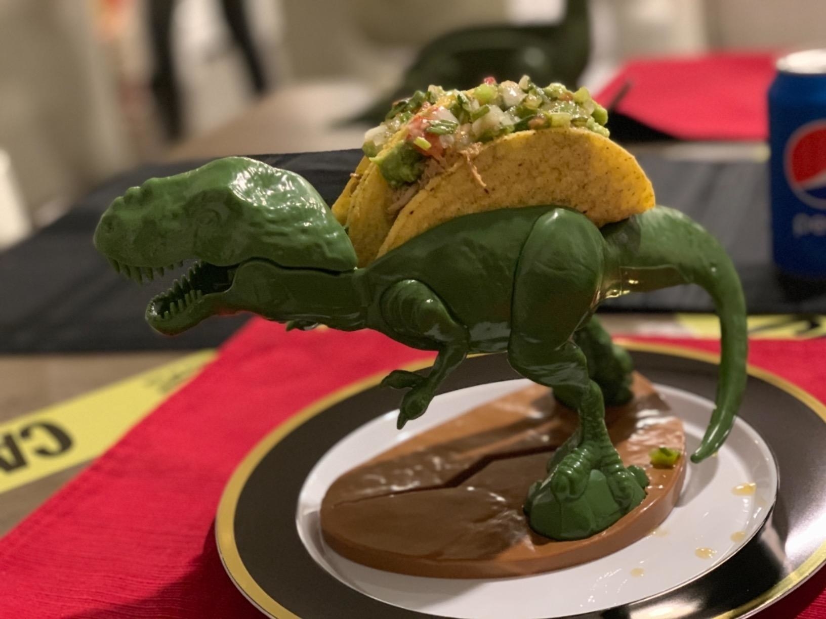 green t rex figure holding two hard shell tacos on his back