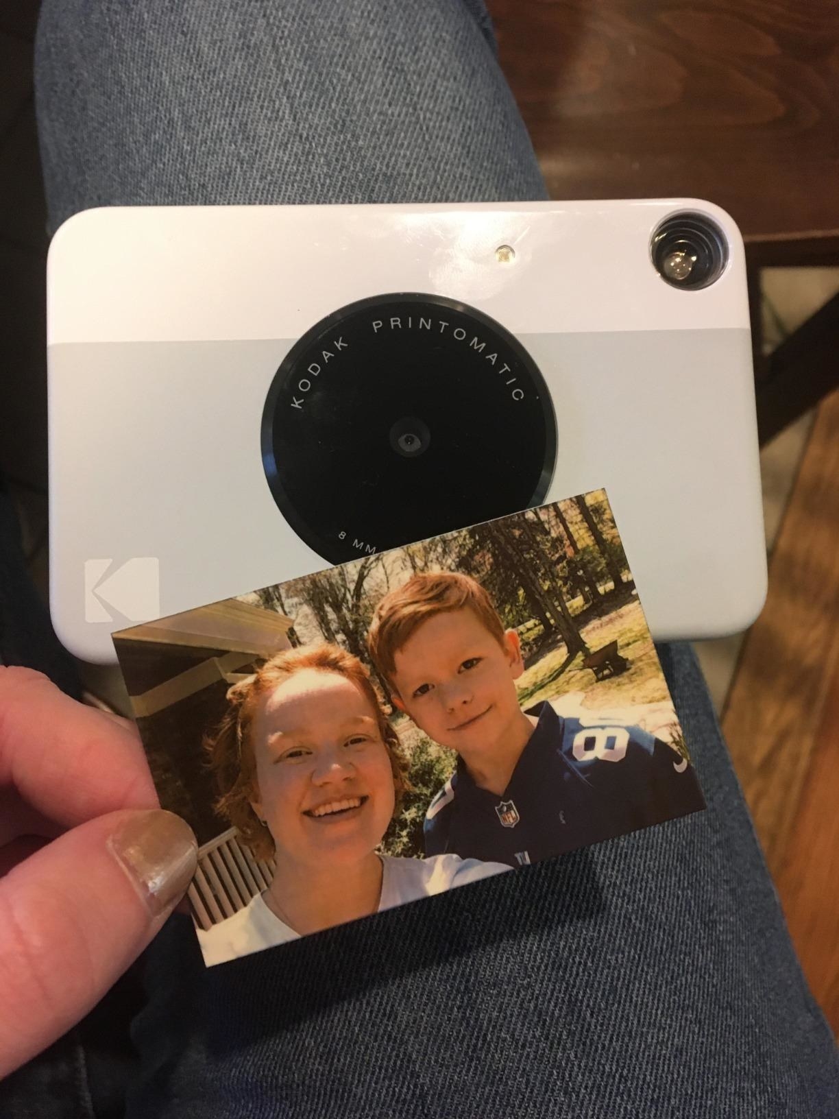 reviewer showing the white instant print camera with a photo of two people printed from the camera