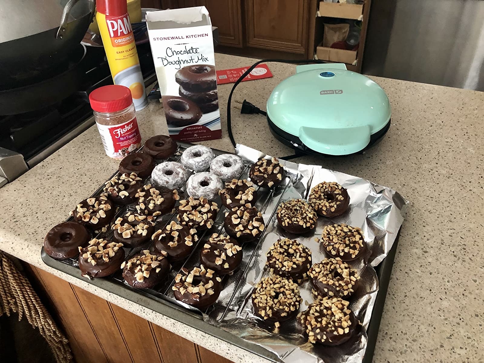 the mini donut maker on a counter with a pan full of mini chocolate donuts in front of it cover in nuts and powdered sugar and all the ingredients on the counter behind the pan
