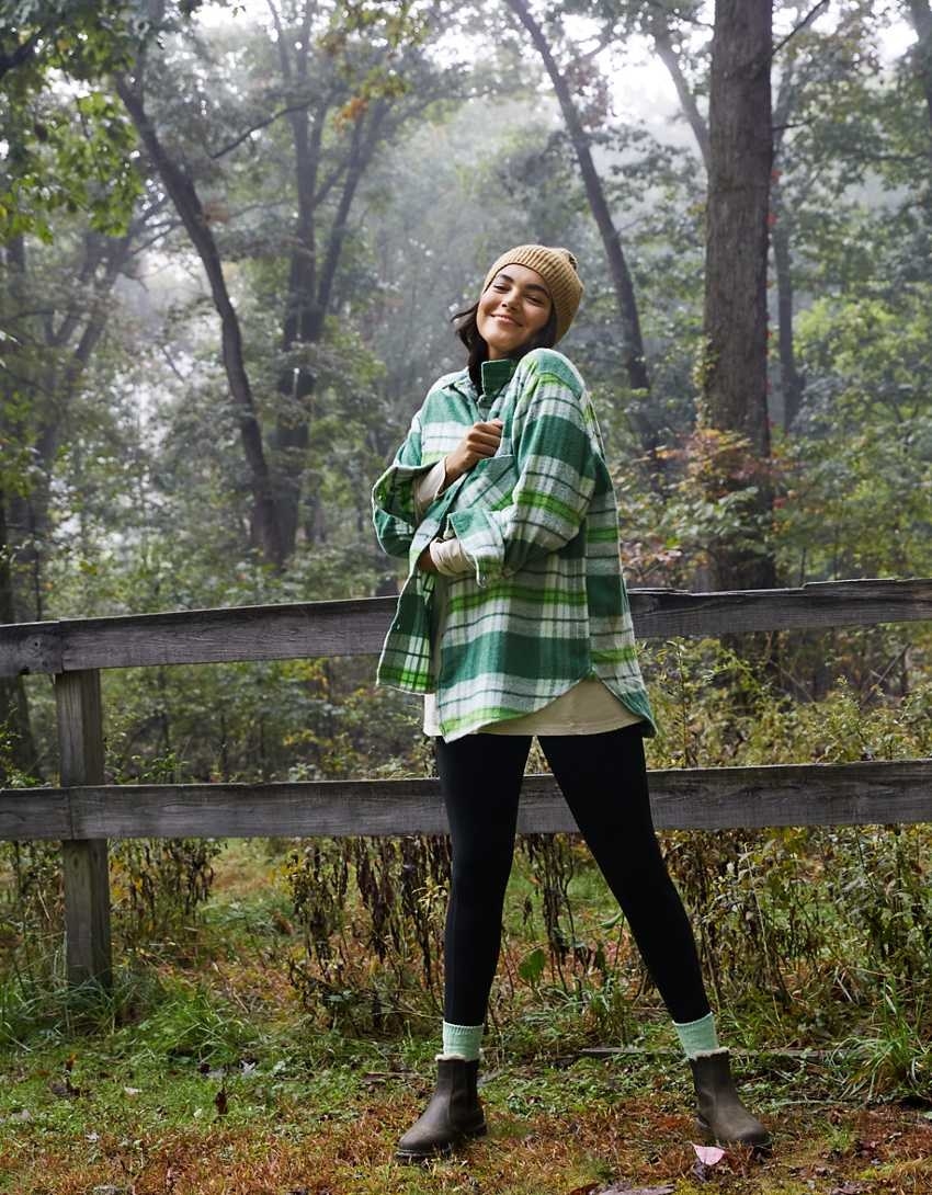 model wearing the green and white plaid shirt with black pants, green socks, and brown boots, standing in a foresty area in front of a wooden fence