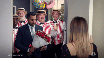 Kal Penn as Derrick Modi stands in a doorway with a barbershop quartet and balloons behind him as he hands a bouquet of roses to the women holding the door open in &quot;Sunnyside&quot;