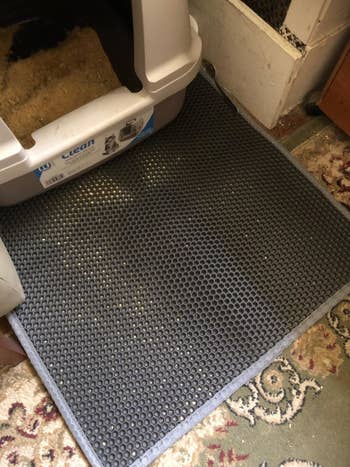 A reviewer's photo of the trapper mat set up under their litter box on a carpet floor, with litter visible trapped in the honeycomb