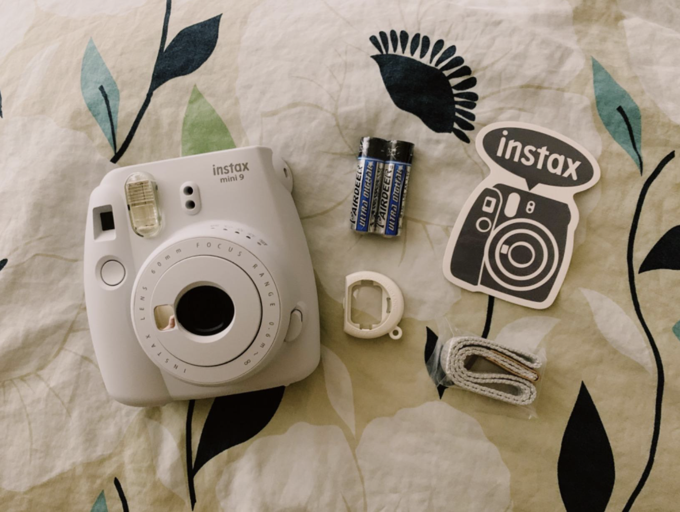 the Instax camera in white with batteries, camera and strap displayed on a bedspread