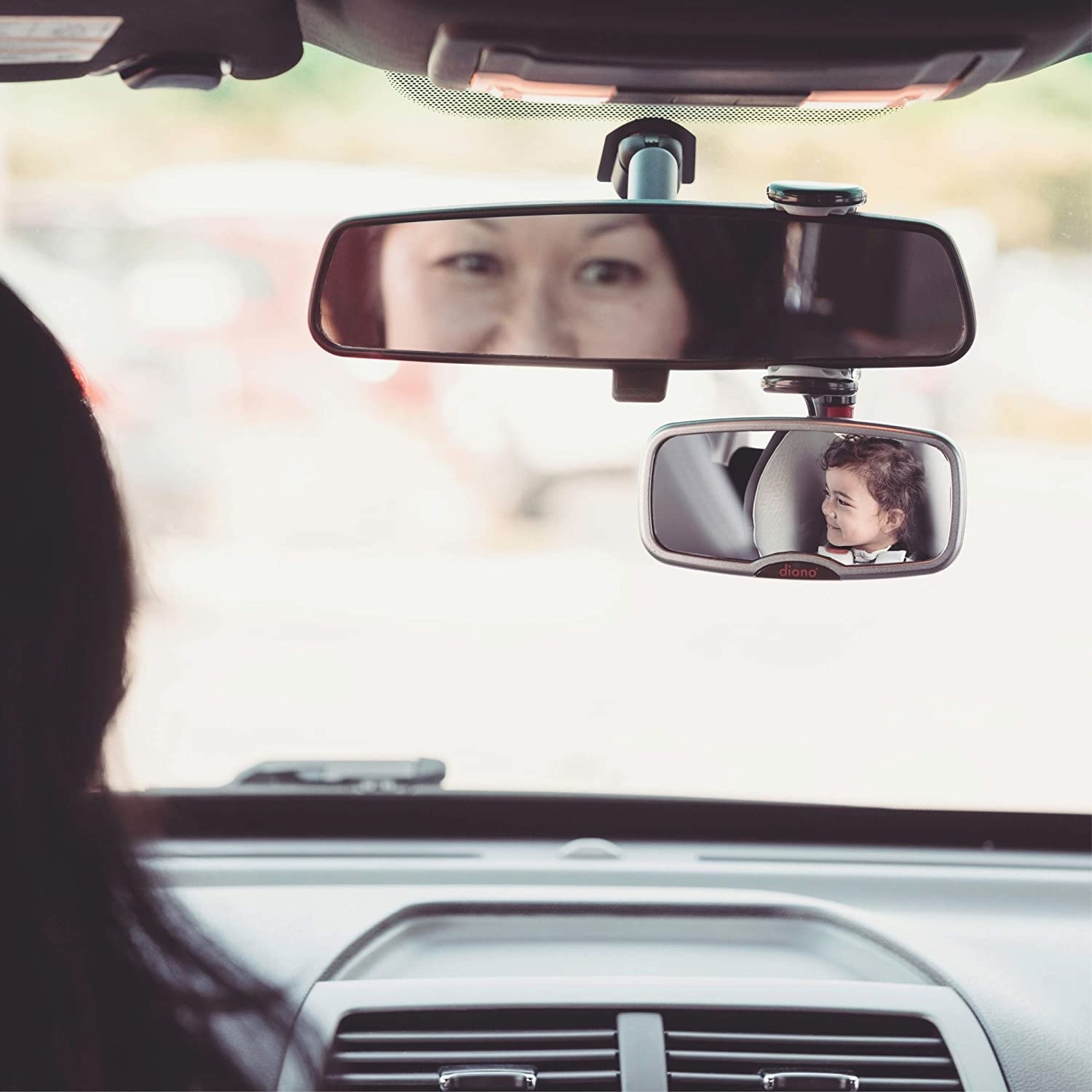 a car&#x27;s rearview mirror with the silver smaller mirror hanging underneath it and you can see a child sitting in the backseat
