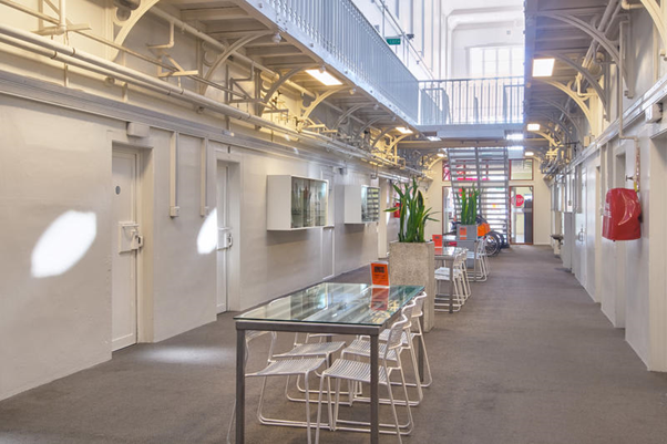 Hostel common area in old jailhouse. Common area has tables and chairs with tall concrete planters to match the light walls, metal stairs, and upper level walkway to the jail cell rooms.