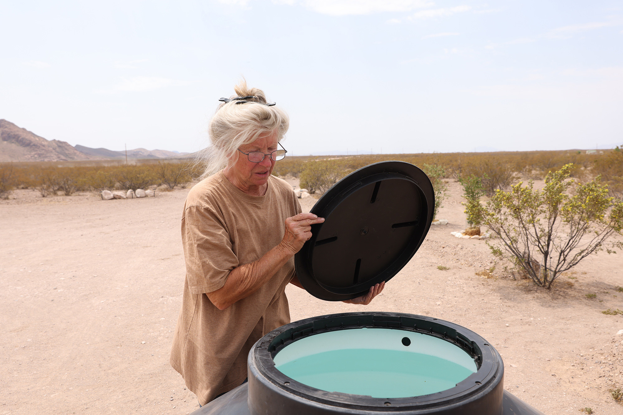 A woman with white hair in a T-shirt looks into a water well in a dry desert landscape