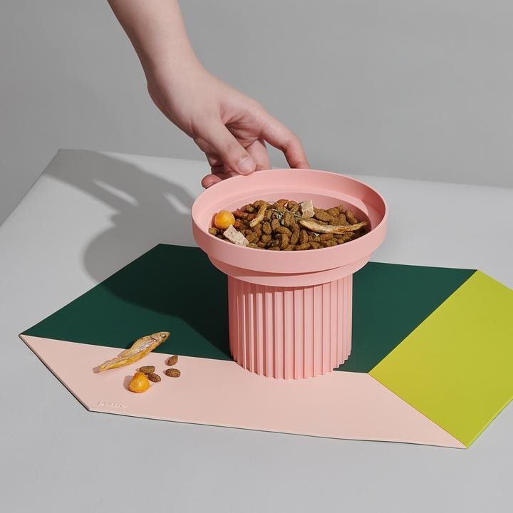 A green, yellow, and light pink geometric feeding mat with a bowl of cat food being set on top of it