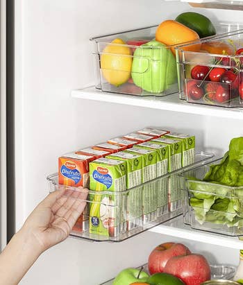 A model pulling the handle of the clear organizer in a fridge filled with fruit, vegetables, and juice boxes