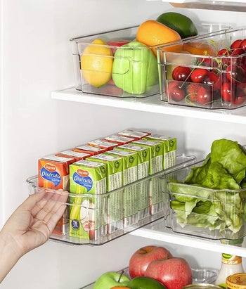 A model pulling the handle of the clear organizer in a fridge filled with fruit, vegetables, and juice boxes