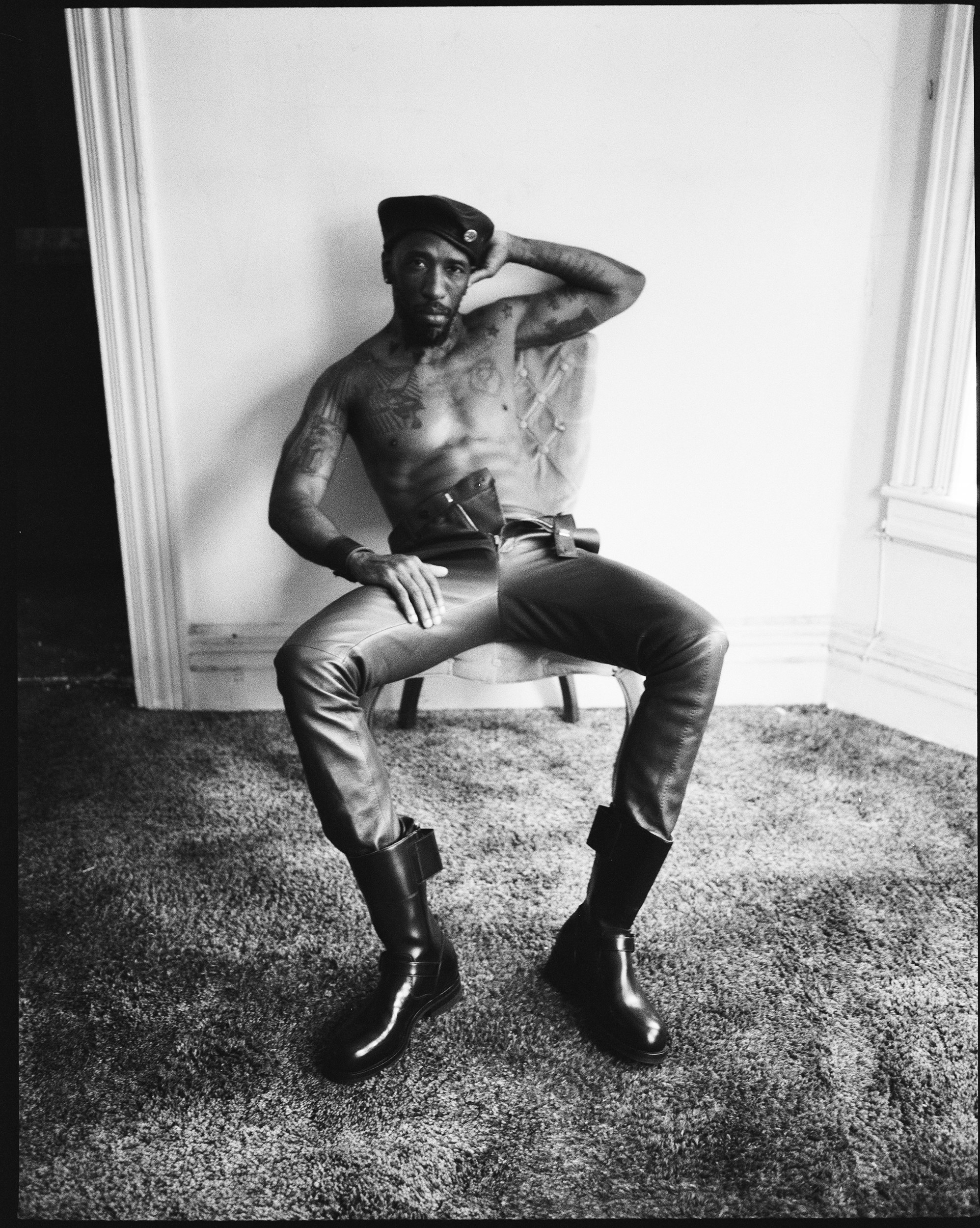 A shirtless man in leather pants and leather boots sitting in a chair in an empty room 