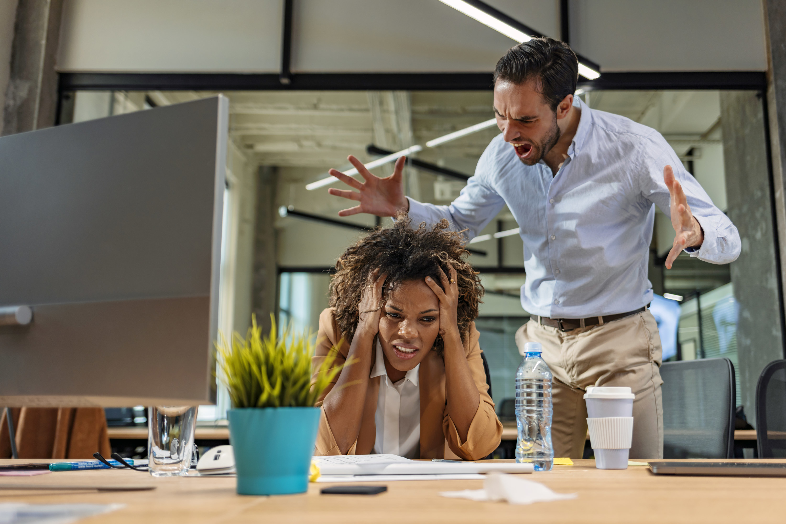 A man standing while yelling at a woman who is sitting at her desk looking stressed