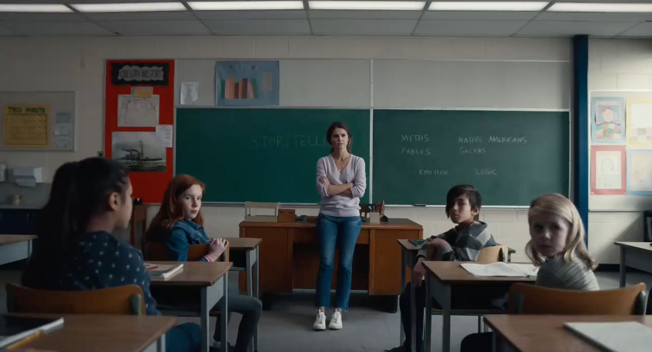 Julia leaning on her desk, standing at the front of the classroom, arms crossed