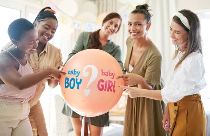 A group of feminine-presenting people pointing to a balloon that says &quot;baby BOY&quot; and &quot;baby GIRL&quot; with a question mark in the middle