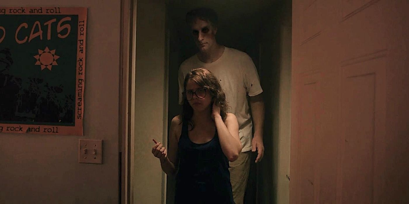 A tall man with hallowed out eye holes lurks behind a woman in a doorway
