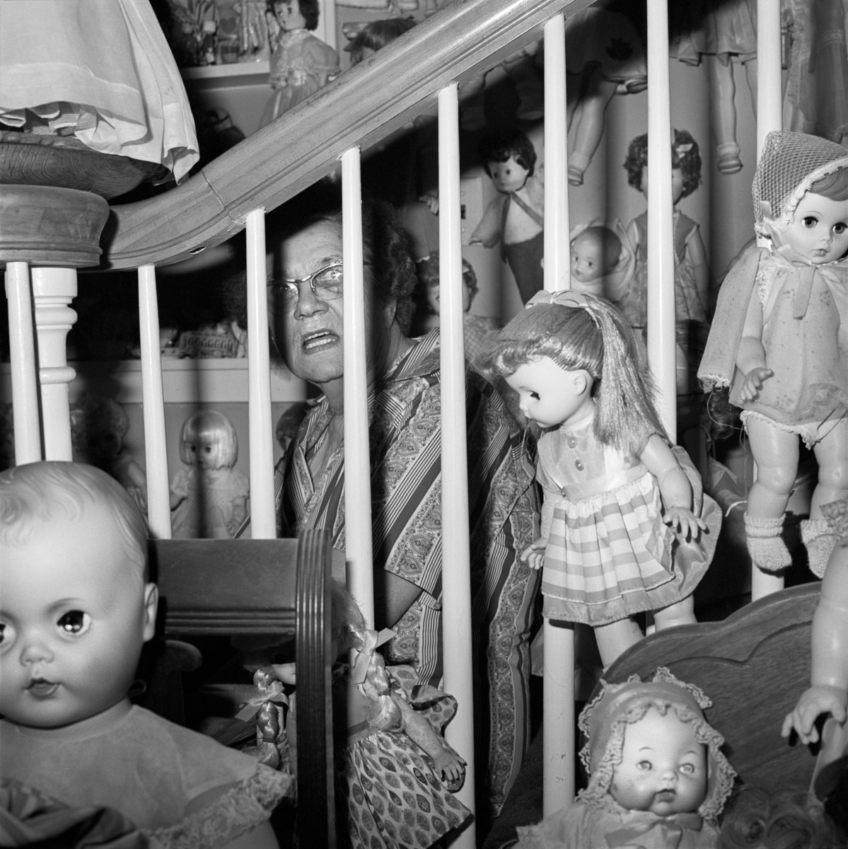 An older woman wearing glasses sits on the stairs behind a railing, surrounded by dolls