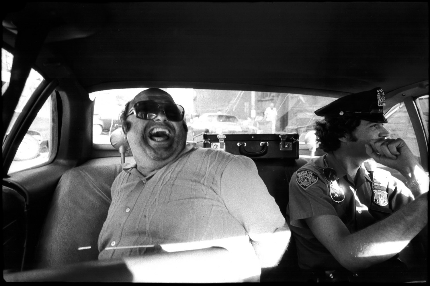 A man laughing in the back of a police car with a cop driving 