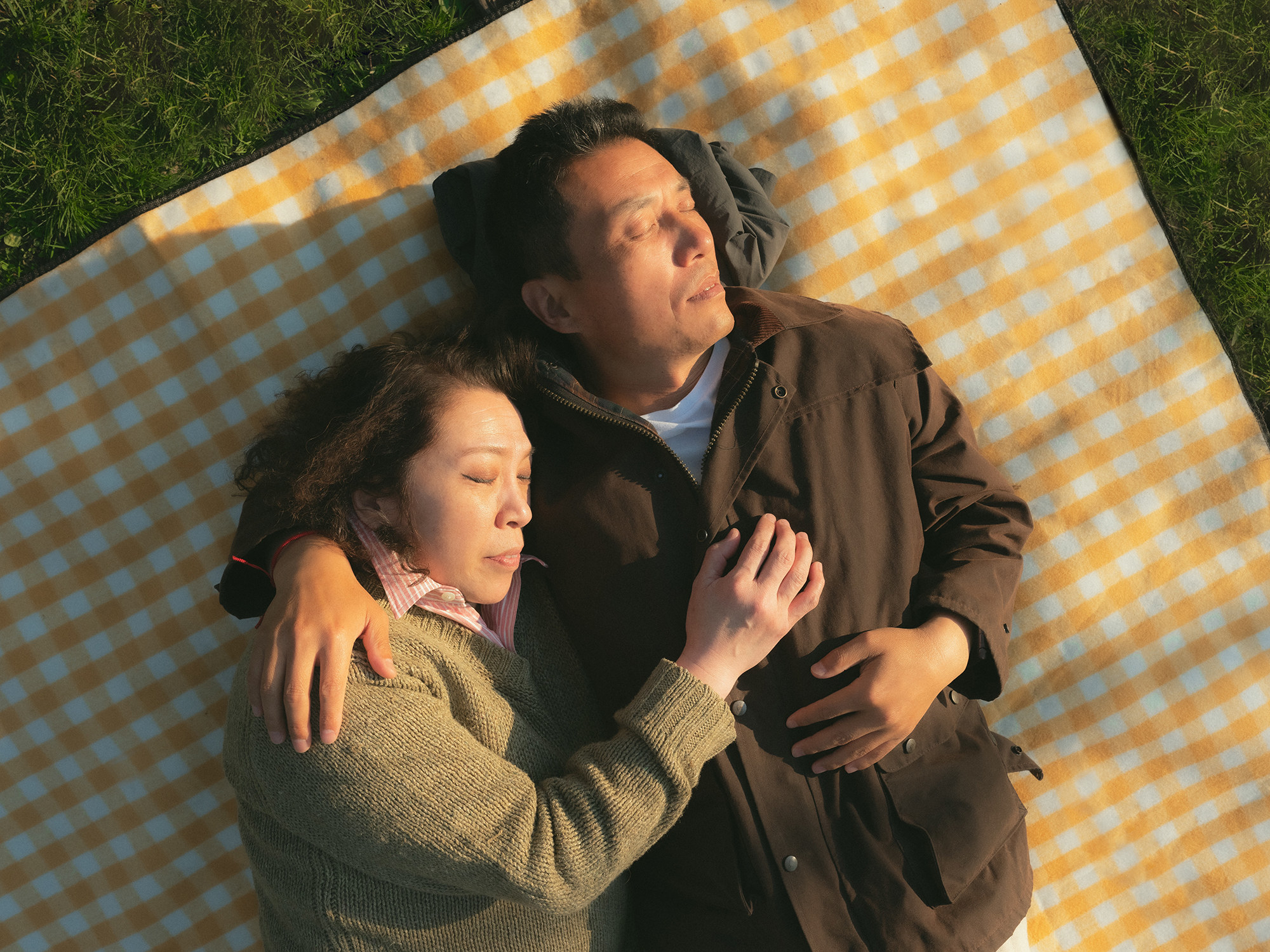 Two older people lying down embracing with their eyes closed on a blanket outside 