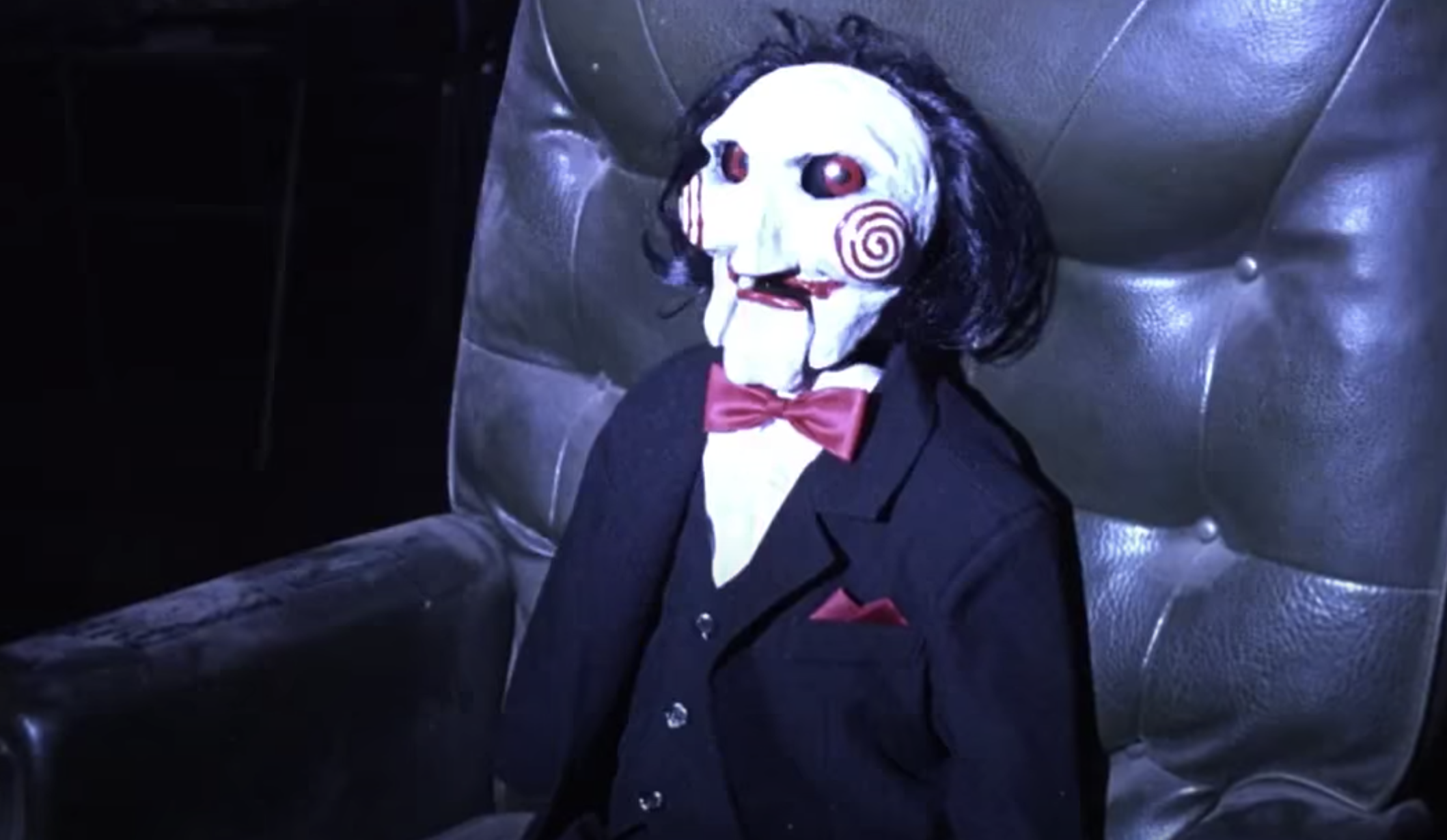 A scary doll with a white face, black hair, and red eyes sits in a chair