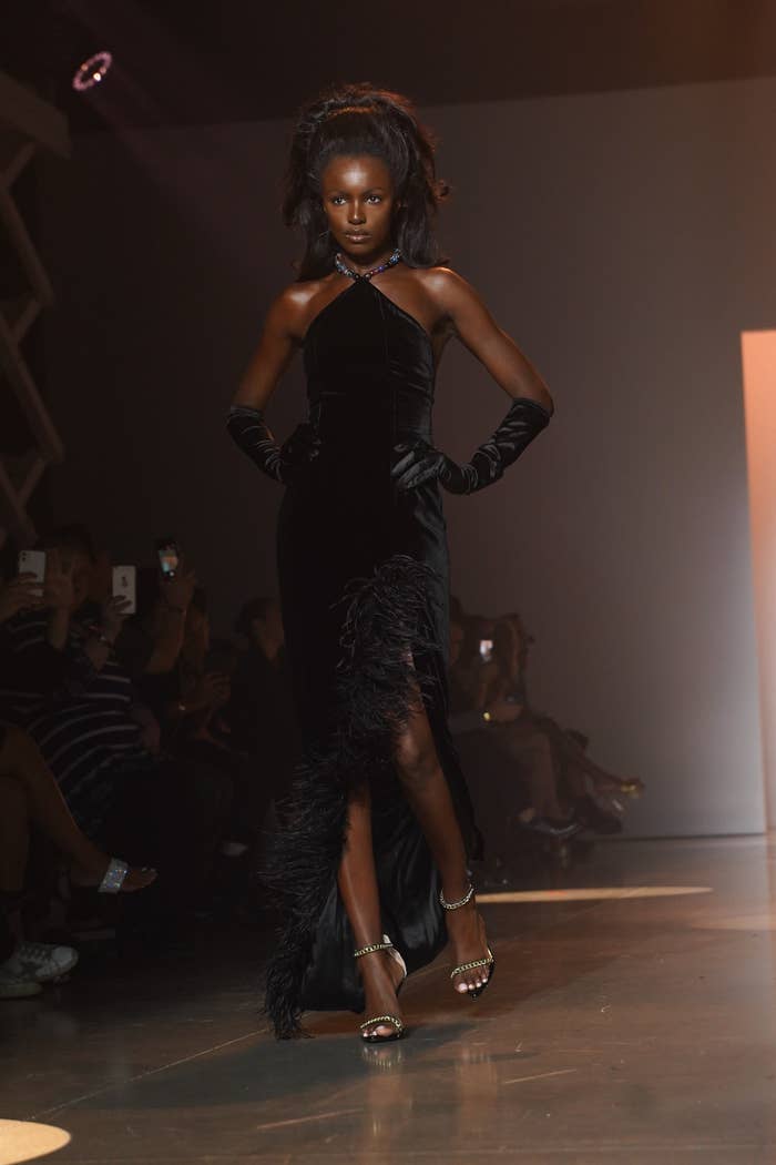 A Black model dressed all in black walks down a runway with her hands on her hips.