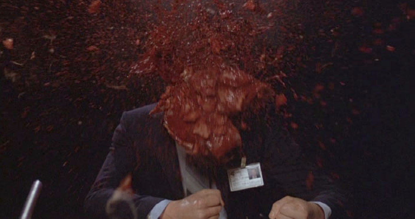 A man is wearing a suit and his head explodes; there is blood and gore everywhere as it flies around