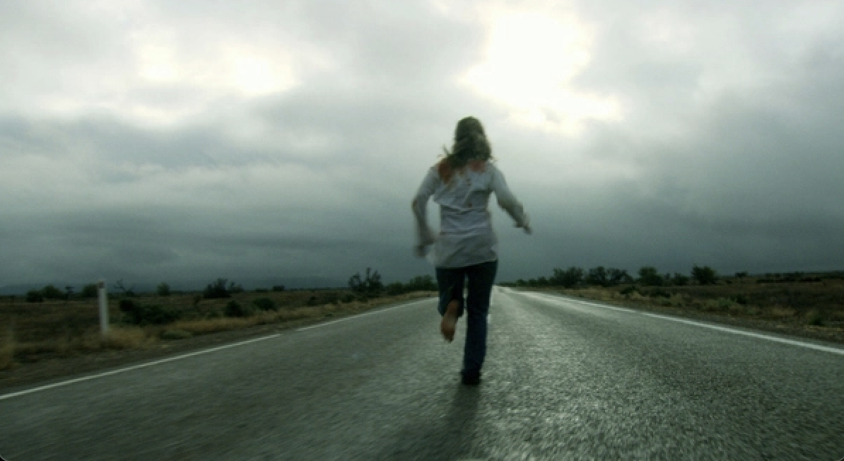 A lone woman runs barefoot and covered in blood on an empty road in the middle of nowhere
