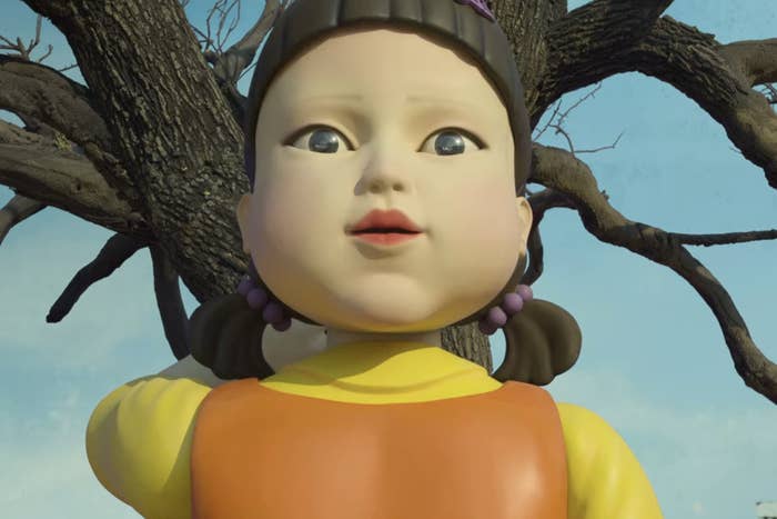 The doll from &quot;Squid Game&quot; wearing a frock with pony tail on each side looking to the camera. There is a tree where the doll is resting her one hand.