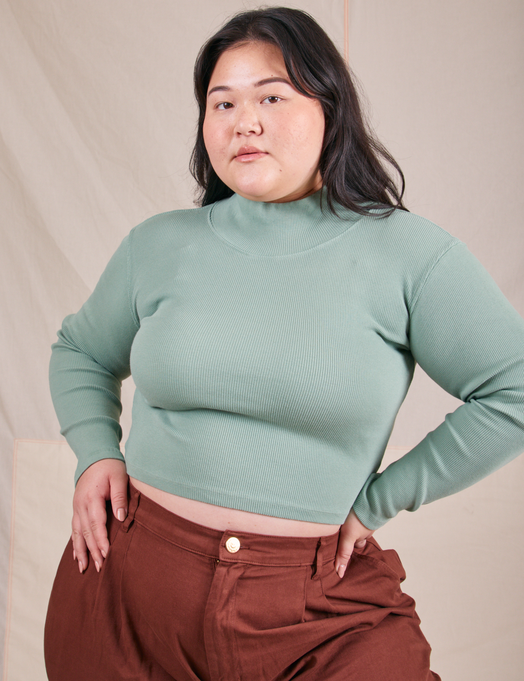 Model is wearing a sage green turtleneck crop sweater and maroon trousers