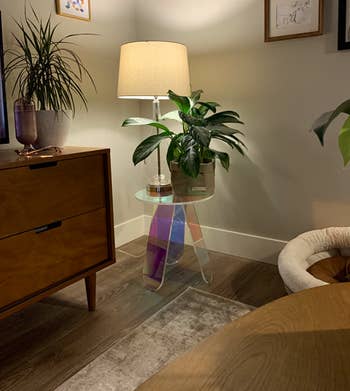 a reviewer's three-legged iridescent acrylic end table with a circular top and a plant sitting on top of it in their living room with a lamp on behind it