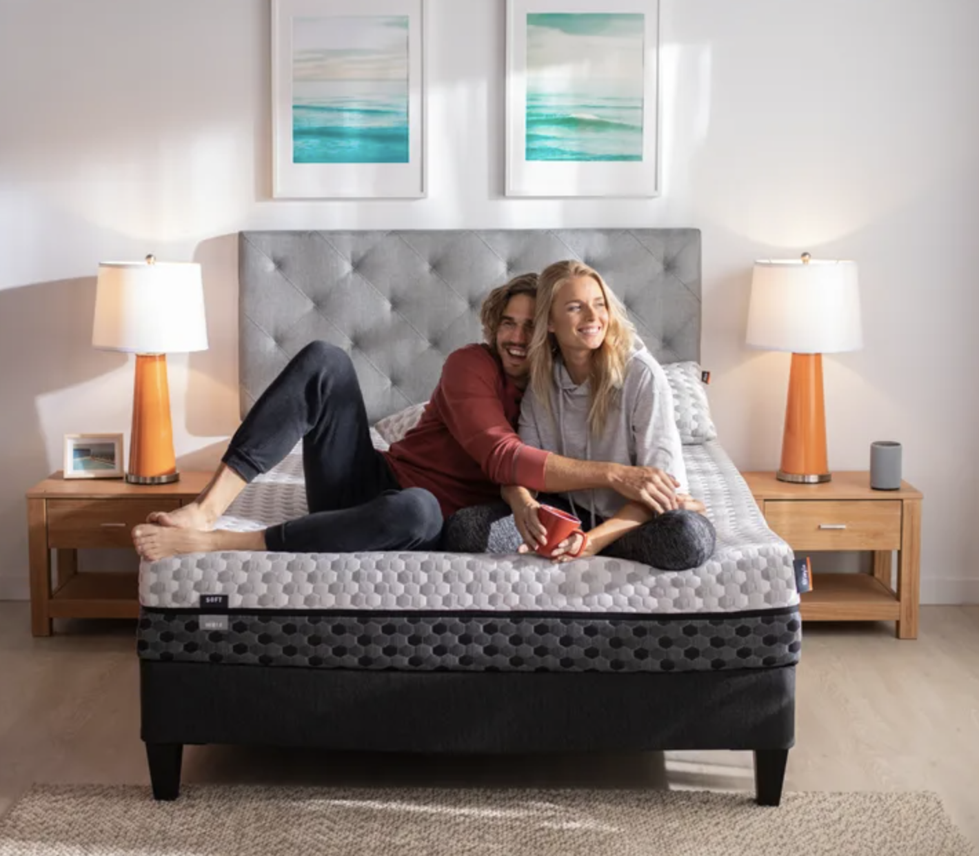 Two models are sitting on the mattress on top of a bed frame