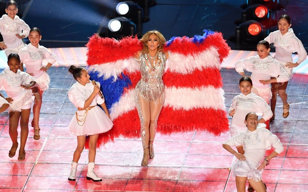 Jlo raising a feathered Puerto Rican flag behind her as she performs with her daughter