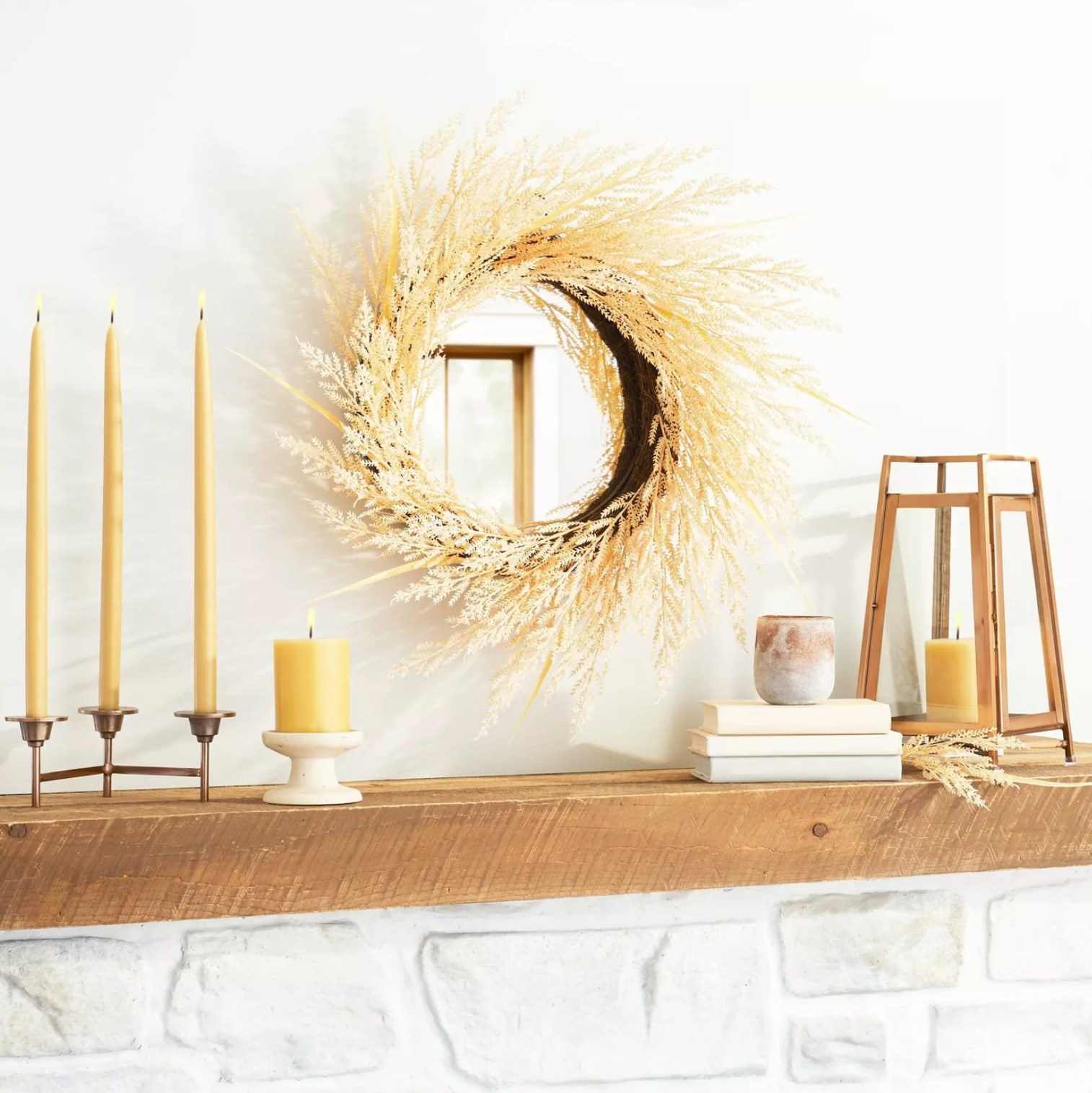 the faux plant wreath hanging around a circle mirror on a wall with candles, books, and other decorations on the wooden hearth beneath it