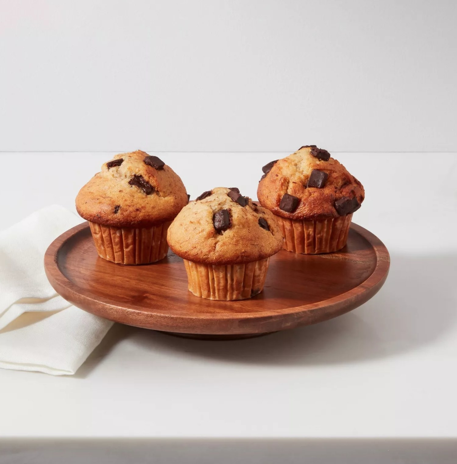 the wooden cake stand with three chocolate chip muffins on it and a white napkin next to it