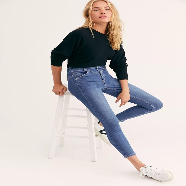 model wearing the blue jeans with white sneakers and a black 3/4 sleeve top sitting on a white stool