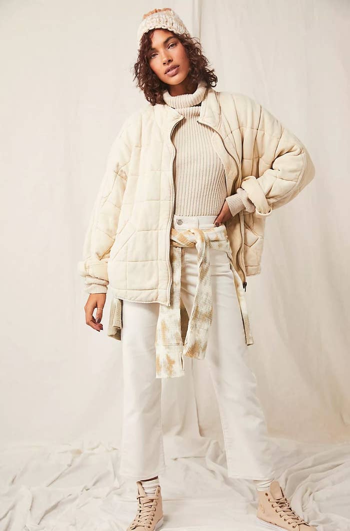 model wearing the cream jacket over white jeans and a tan turtleneck with a tan and white shirt tied around the waist, tan sneakers and a cream knit beanie