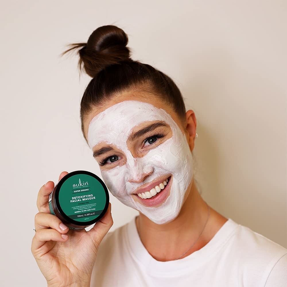 A person holding product in hand with applied clay mask on face