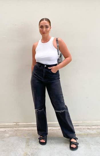 model wearing the black jeans, which have rips on the knee, with a white tank top and black sandals