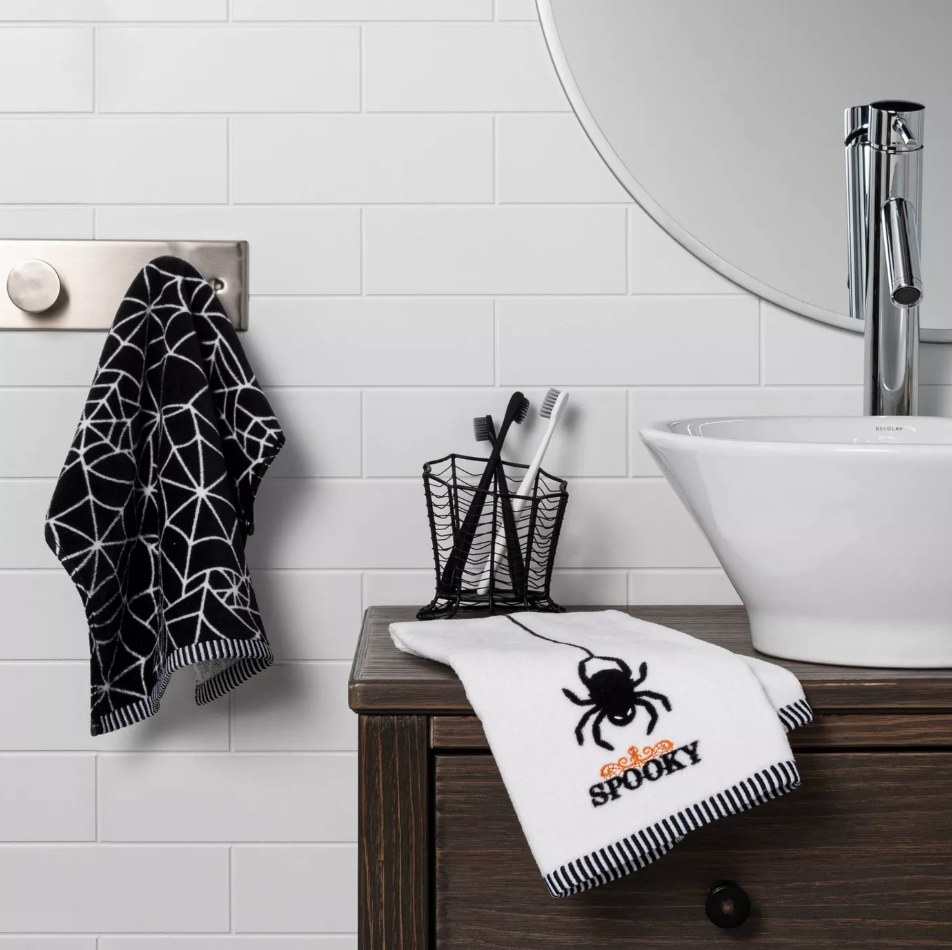 Black hand towel with white cob webs on it hanging from towel rack and white towel with black spider and &quot;spooky&quot; on counter