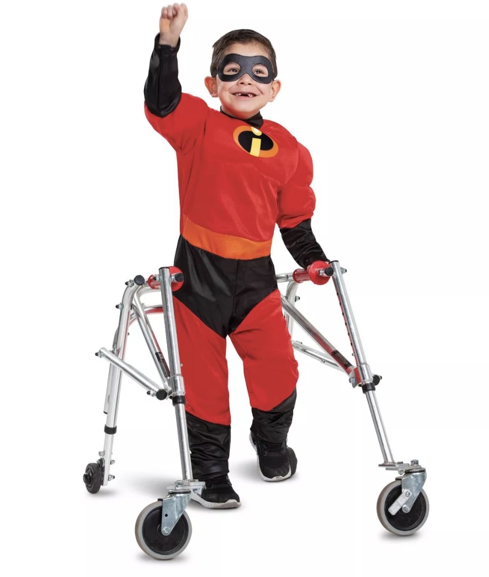 A child standing with an adaptive device wears the black and red incredibles bodysuit with the yellow black and orange &quot;i&quot; logo on the chest