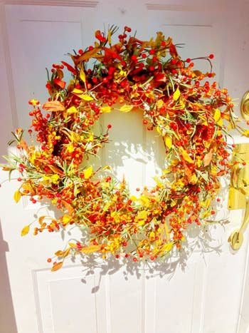 A round wreath with red berries, yellow leaves, and green foliage hanging on a white front door