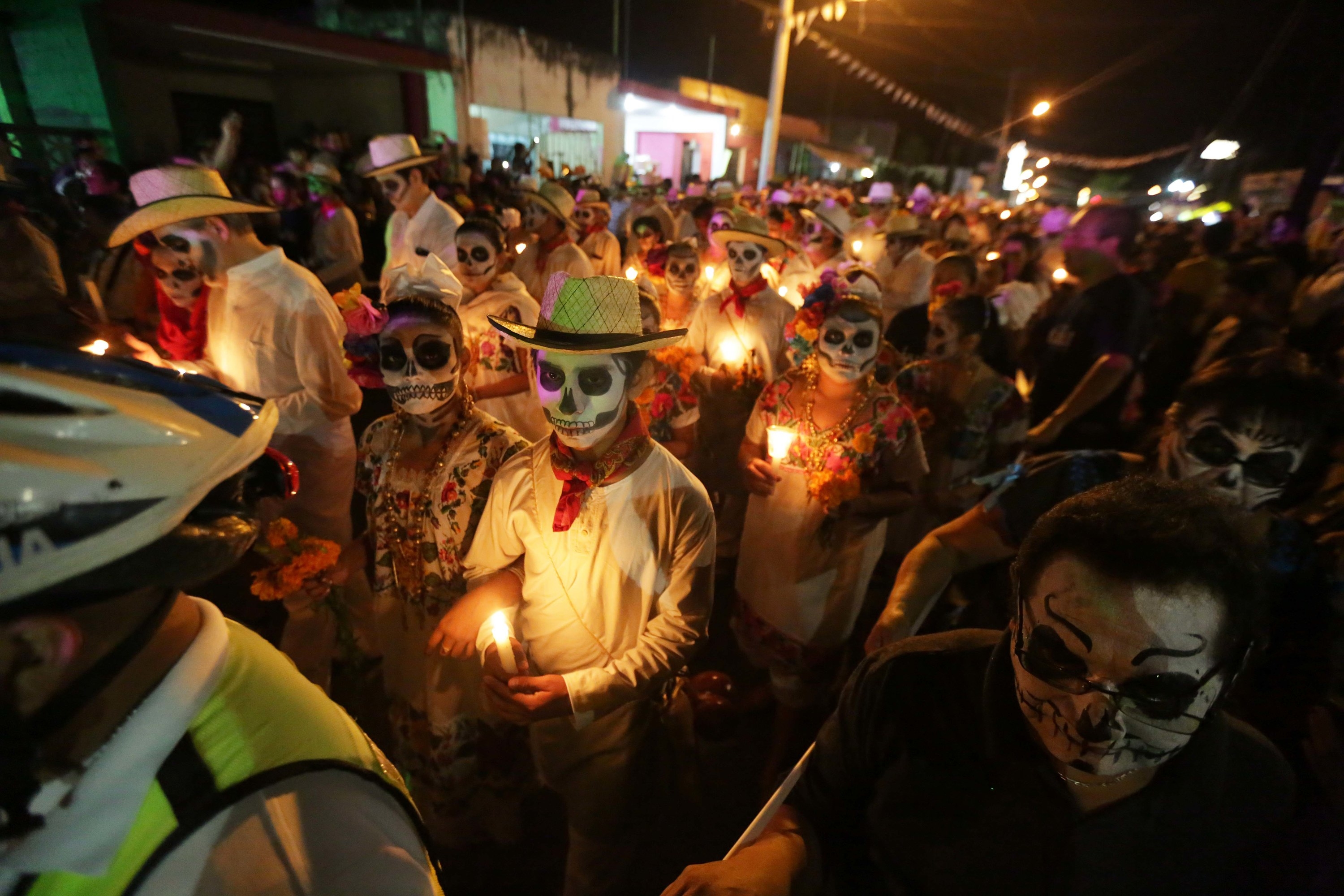 People with painted skull faces and holding candles walk along the street