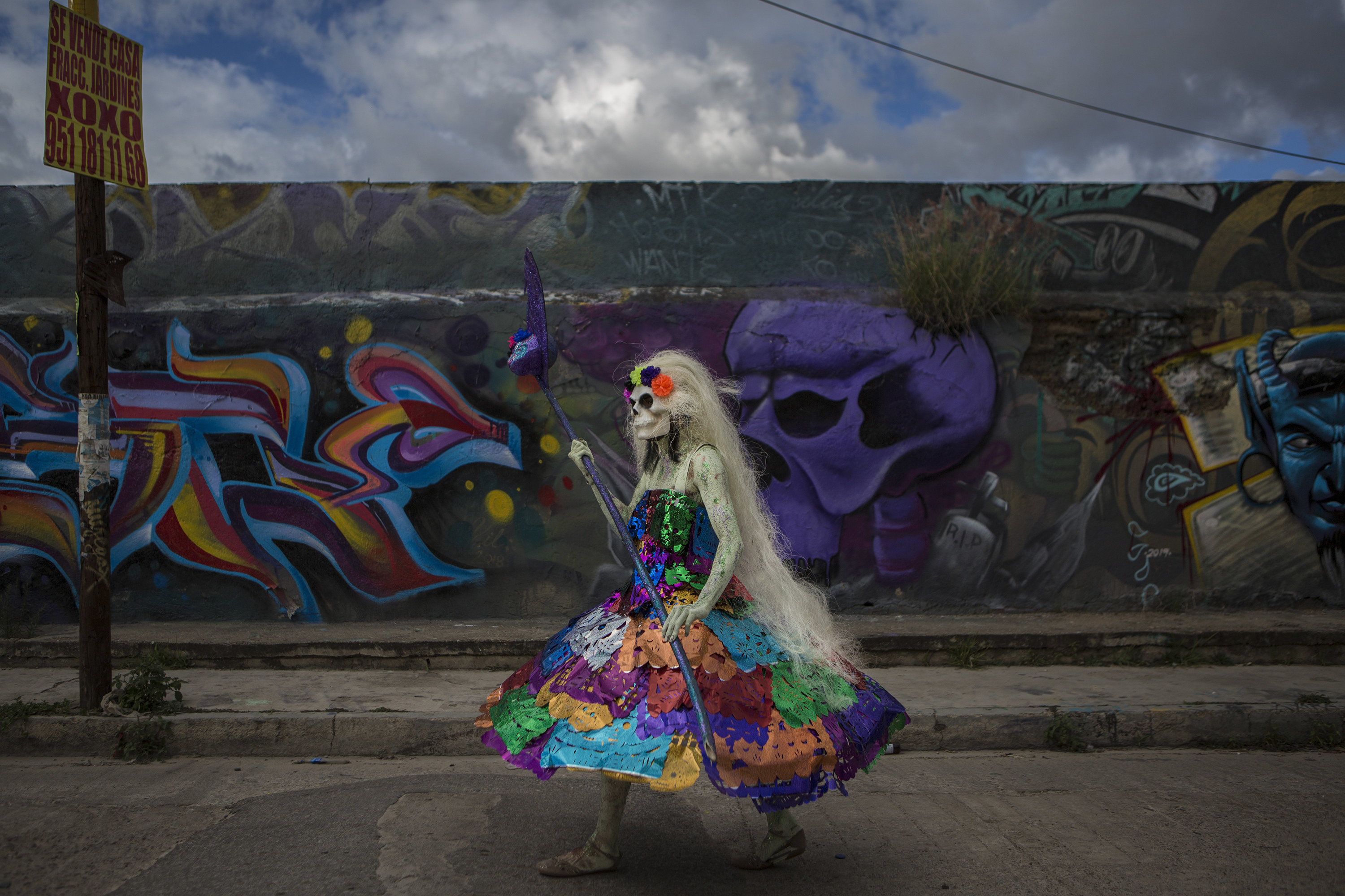 A person wearing a colorful dress, long white wig, and skull face paint walks in front of a wall with graffiti