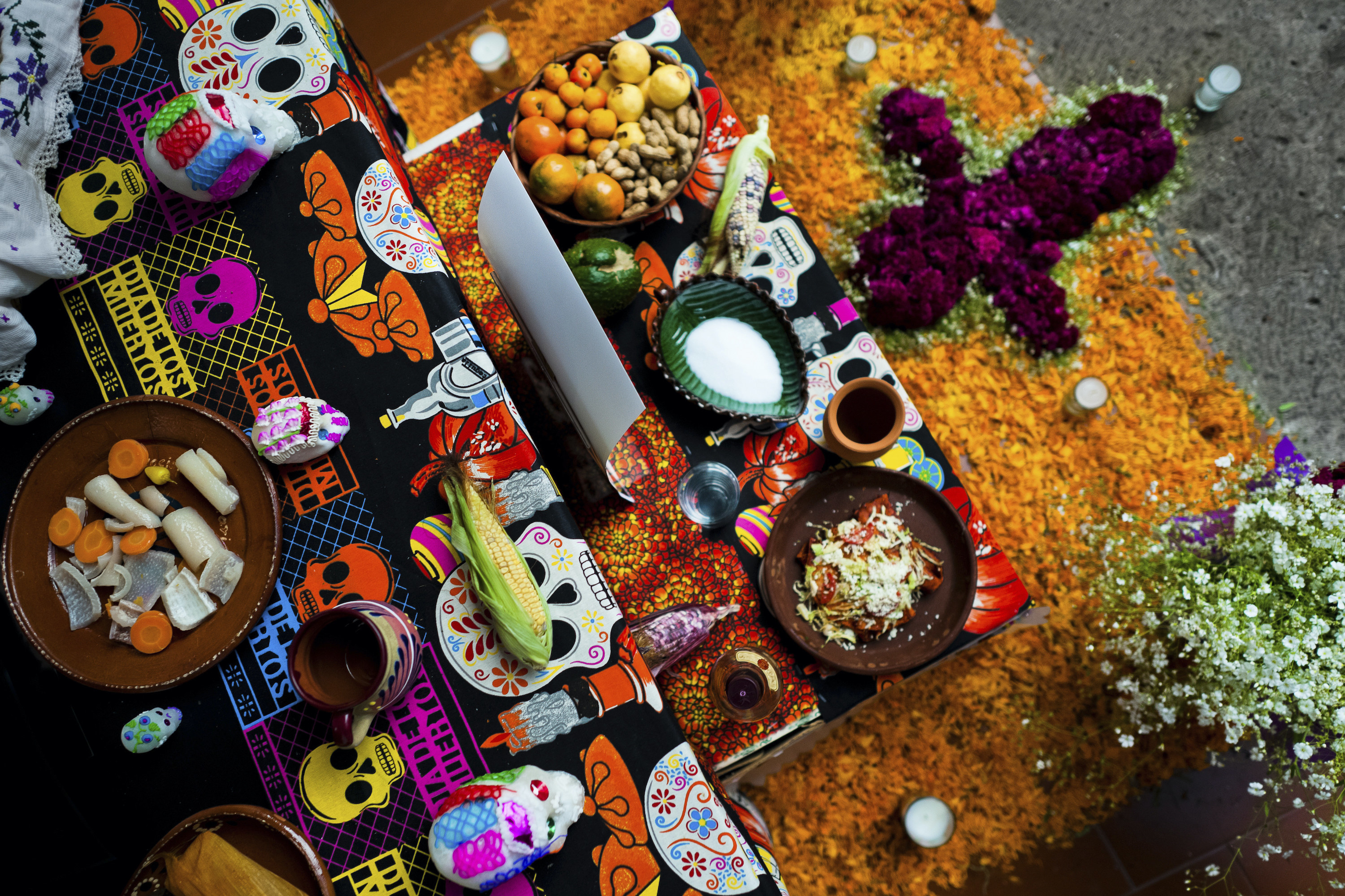 An array of food, including an ear of corn and a bowl of raw vegetables, on a rug and a cloth depicting drawings of skulls and other items