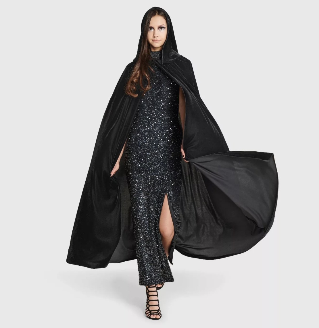An adult in a floor-length black sparkle dress and black sandals wears the large black cape with the hood up