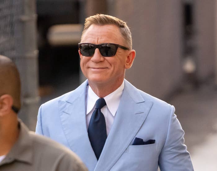 Daniel Craig smiles while wearing a light-colored suit and sunglasses