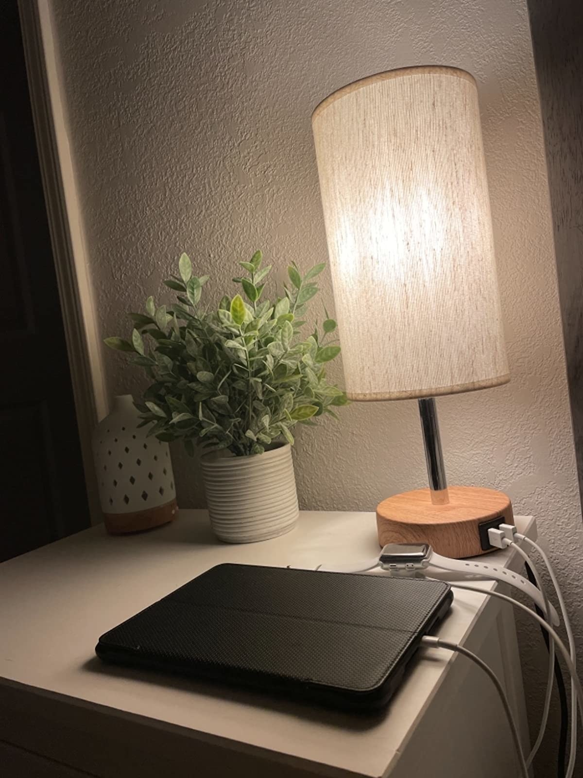 reviewer image of the lamp on a tabletop with a plant and diffuser next to it, and an ipad and apple watch charging in front of it with the cords connected to the two USB ports in the lamp